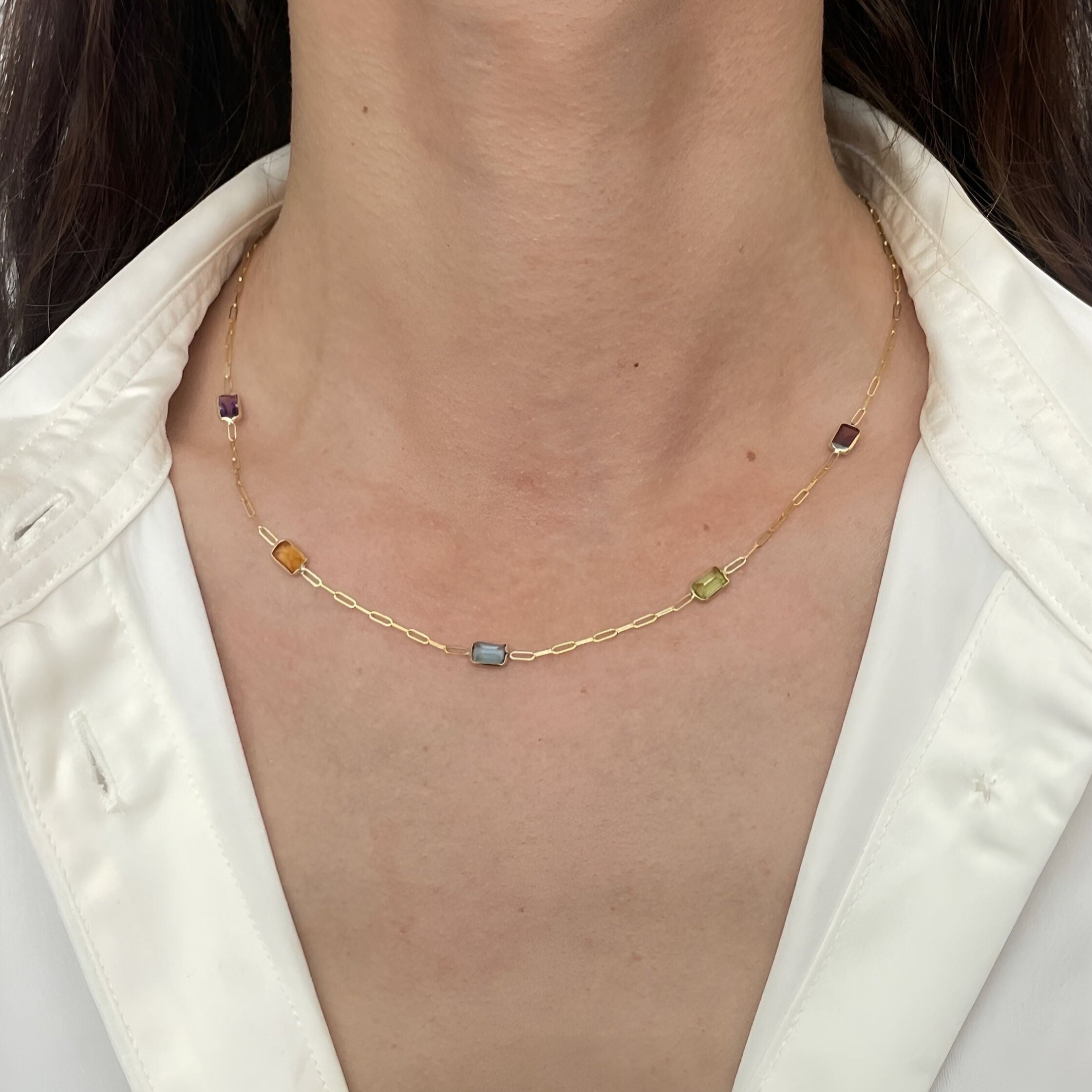 A close-up of a model wearing a delicate gold chain necklace with small, multi-colored gemstone beads, styled with a white satin shirt.