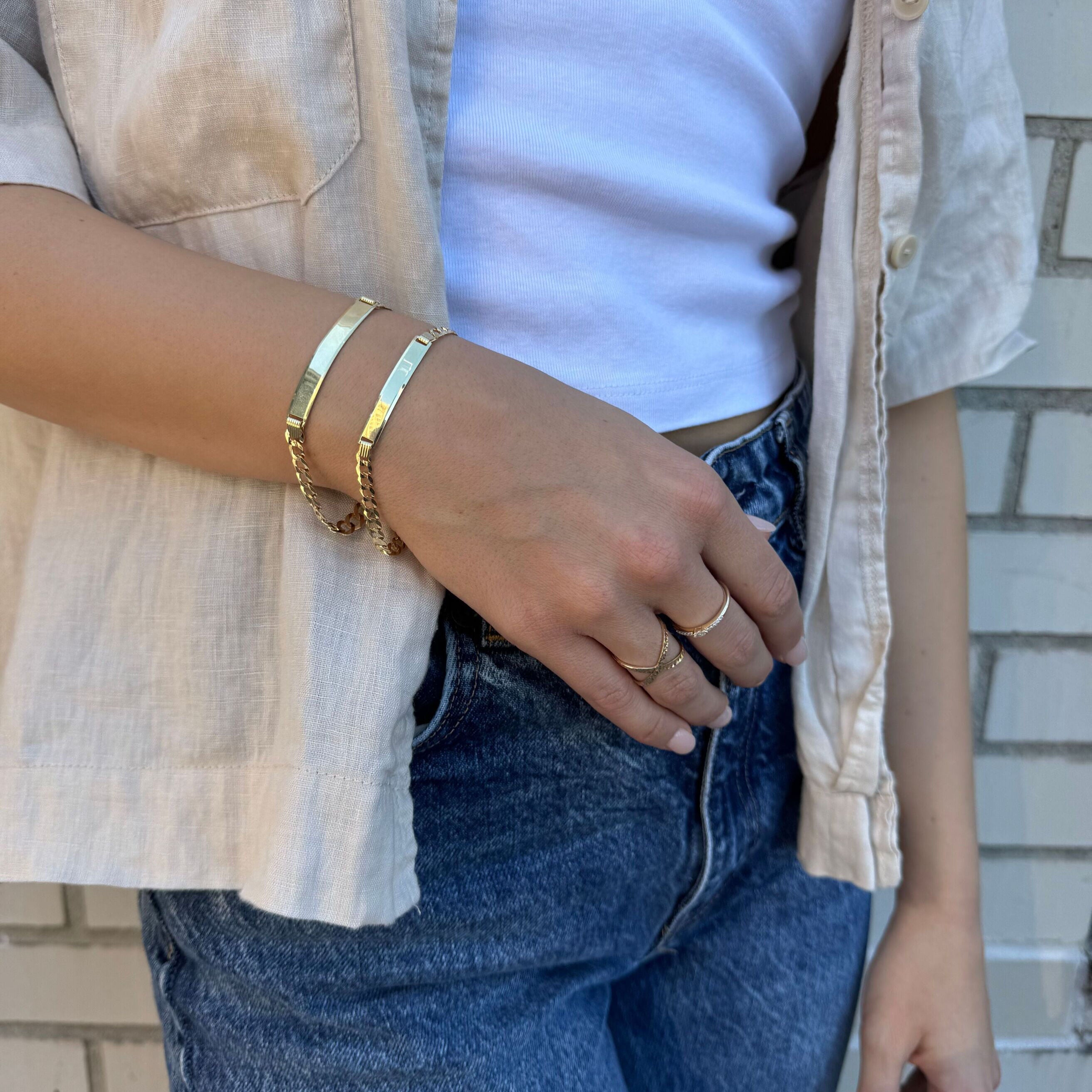 Engravable 14K gold ID bar bracelet stacked with other gold jewelry and styled with relaxed casual clothing. The bracelet’s bold curb links elevate the layered look with timeless elegance, making it suitable for birthdays, anniversary dinners, or casual luxury wear.