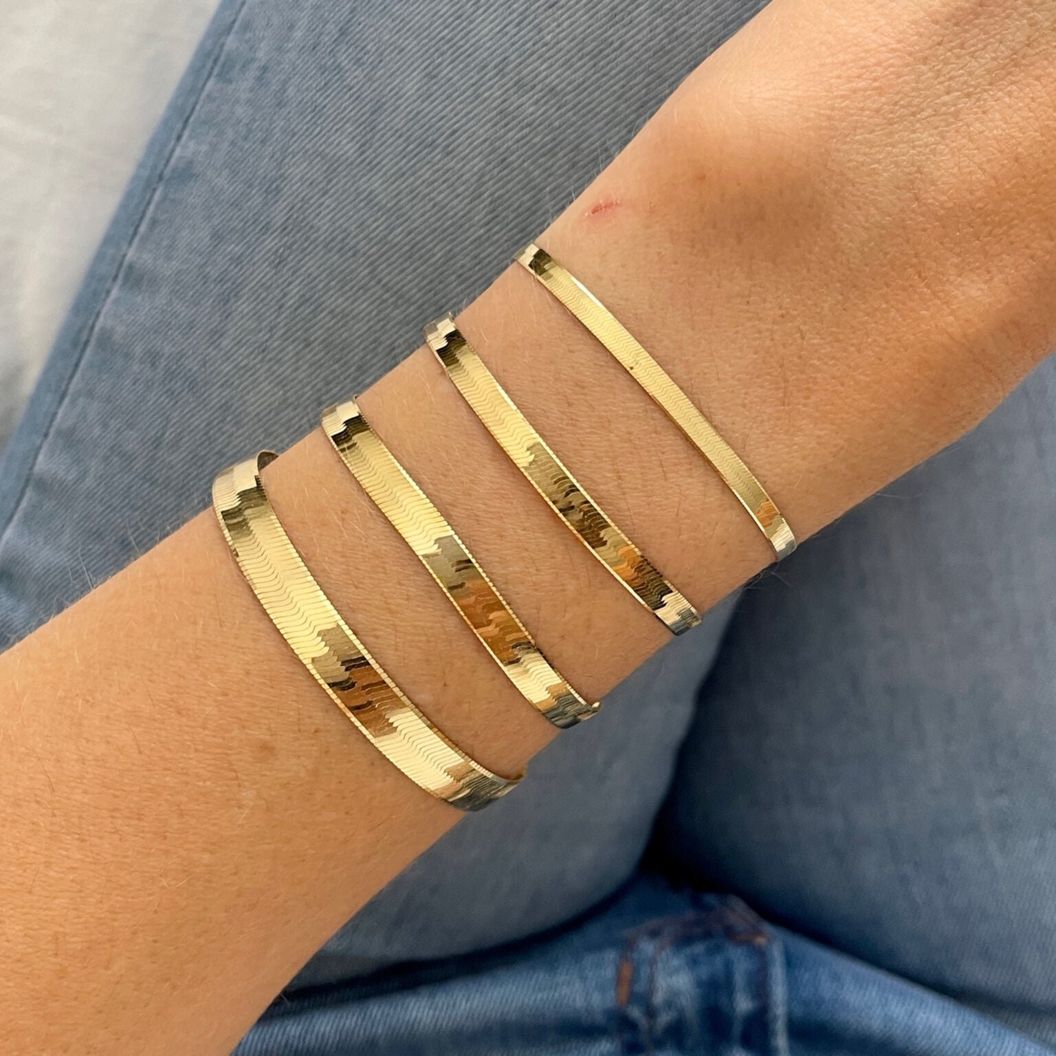 14K yellow gold herringbone bracelets in varied widths styled on the wrist with casual denim jeans. The luminous flat gold links enhance the outfit with a touch of modern elegance and versatility. Perfect for casual meetings, weekend outings, or everyday wear.