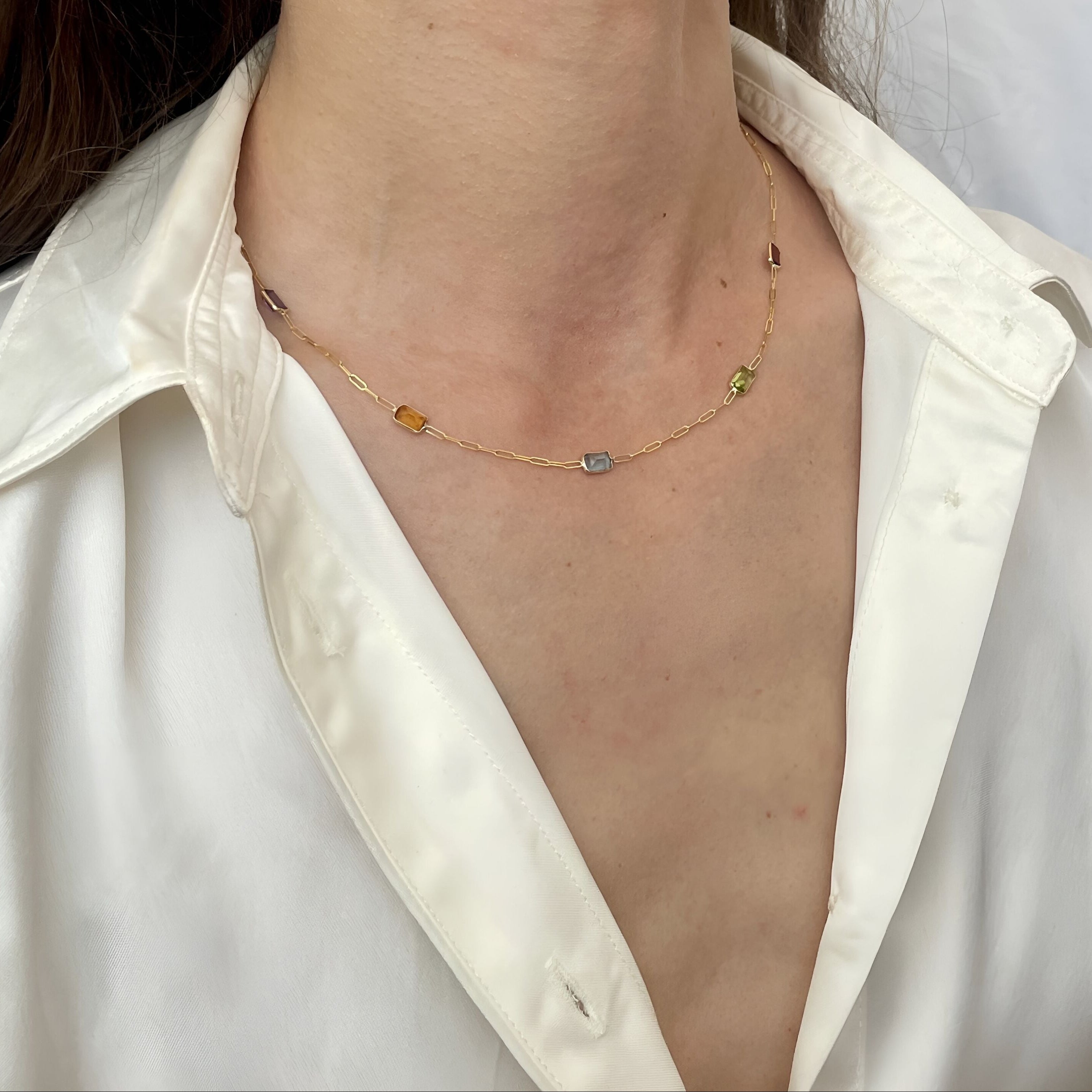 A close-up of a model wearing a delicate gold chain necklace with small, multi-colored gemstone accents, styled with a white satin shirt.