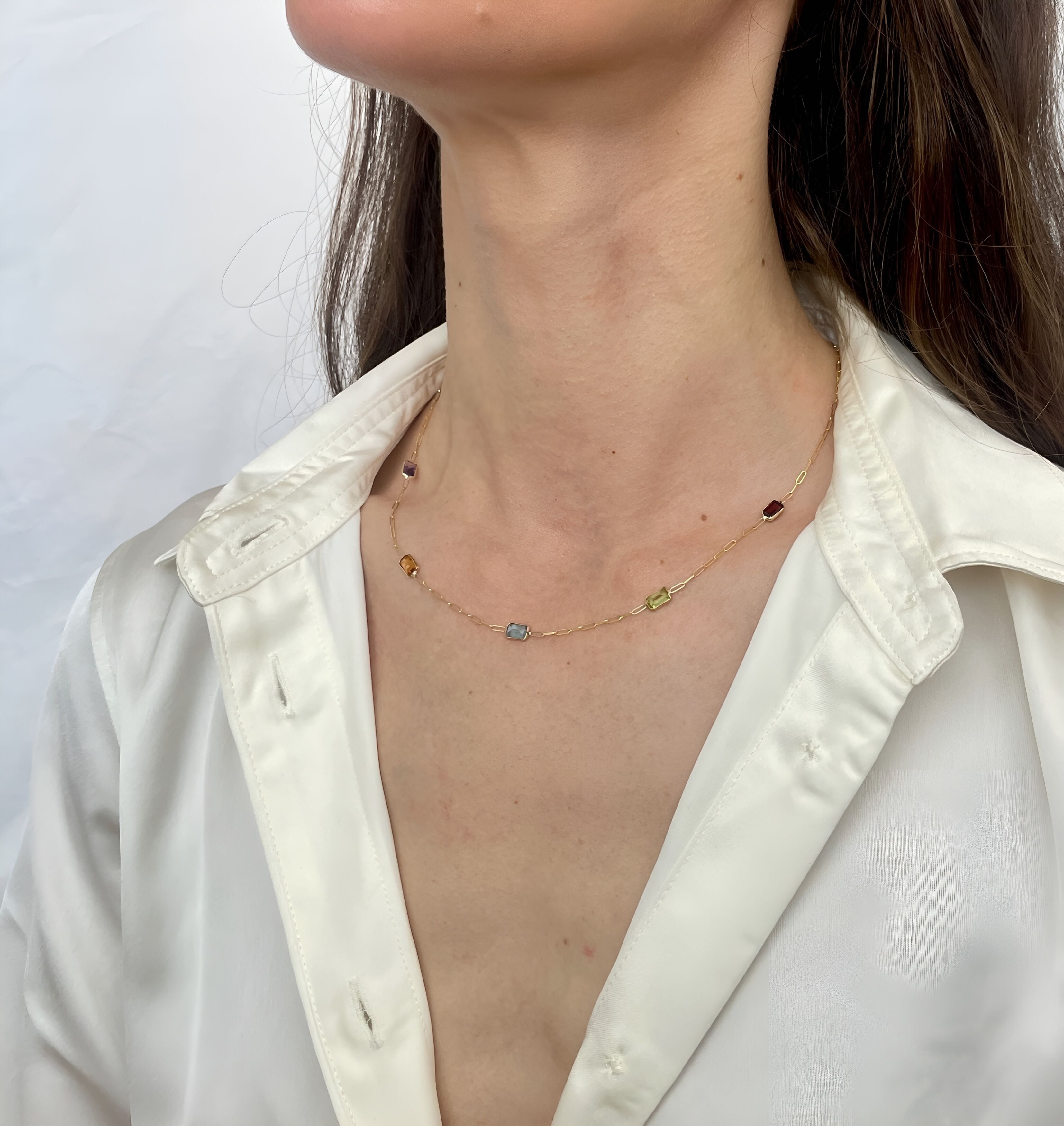 A close-up of a model wearing a delicate gold chain necklace with small, multi-colored gemstone beads, paired with an off-white satin shirt.