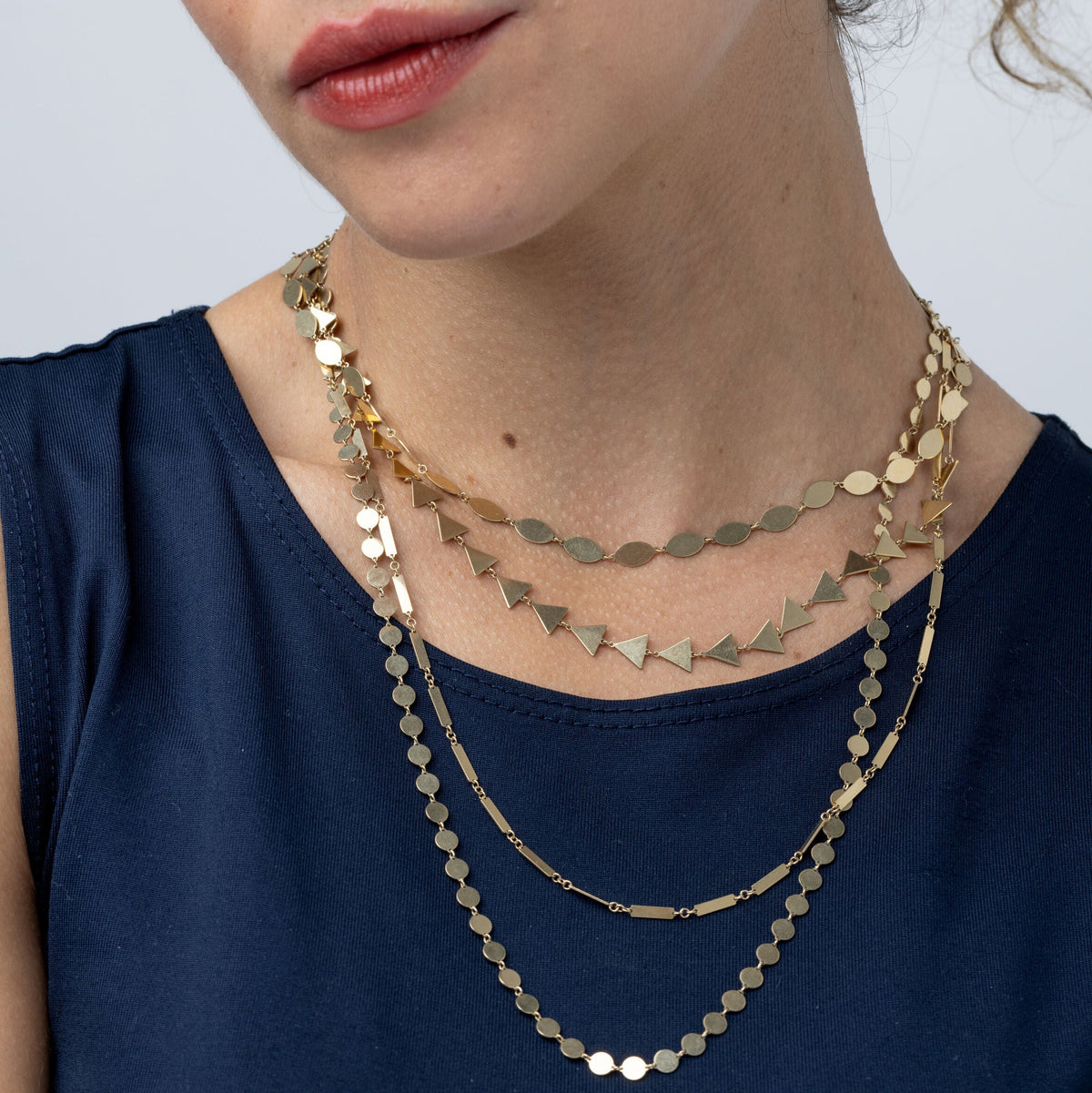 A close-up of a woman wearing a layered gold necklace set with gold disc and triangle charms, paired with a navy blue top.