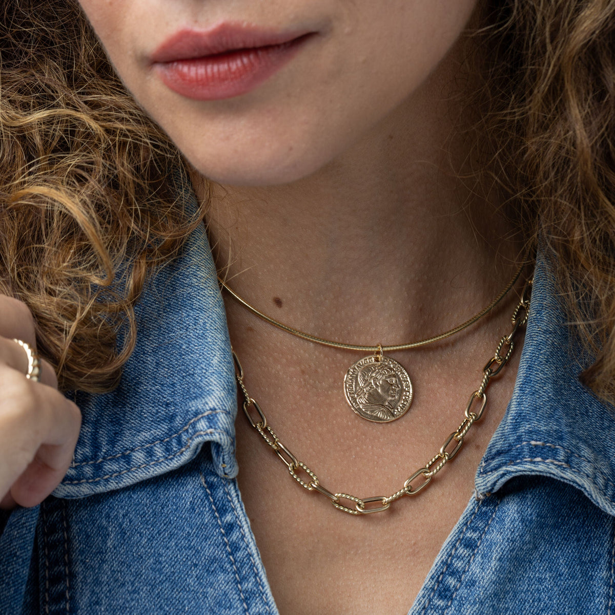 A close-up of a person wearing layered omega gold necklaces, including a thin chain with a round coin pendant and a thicker chain link necklace, paired with a blue denim shirt.