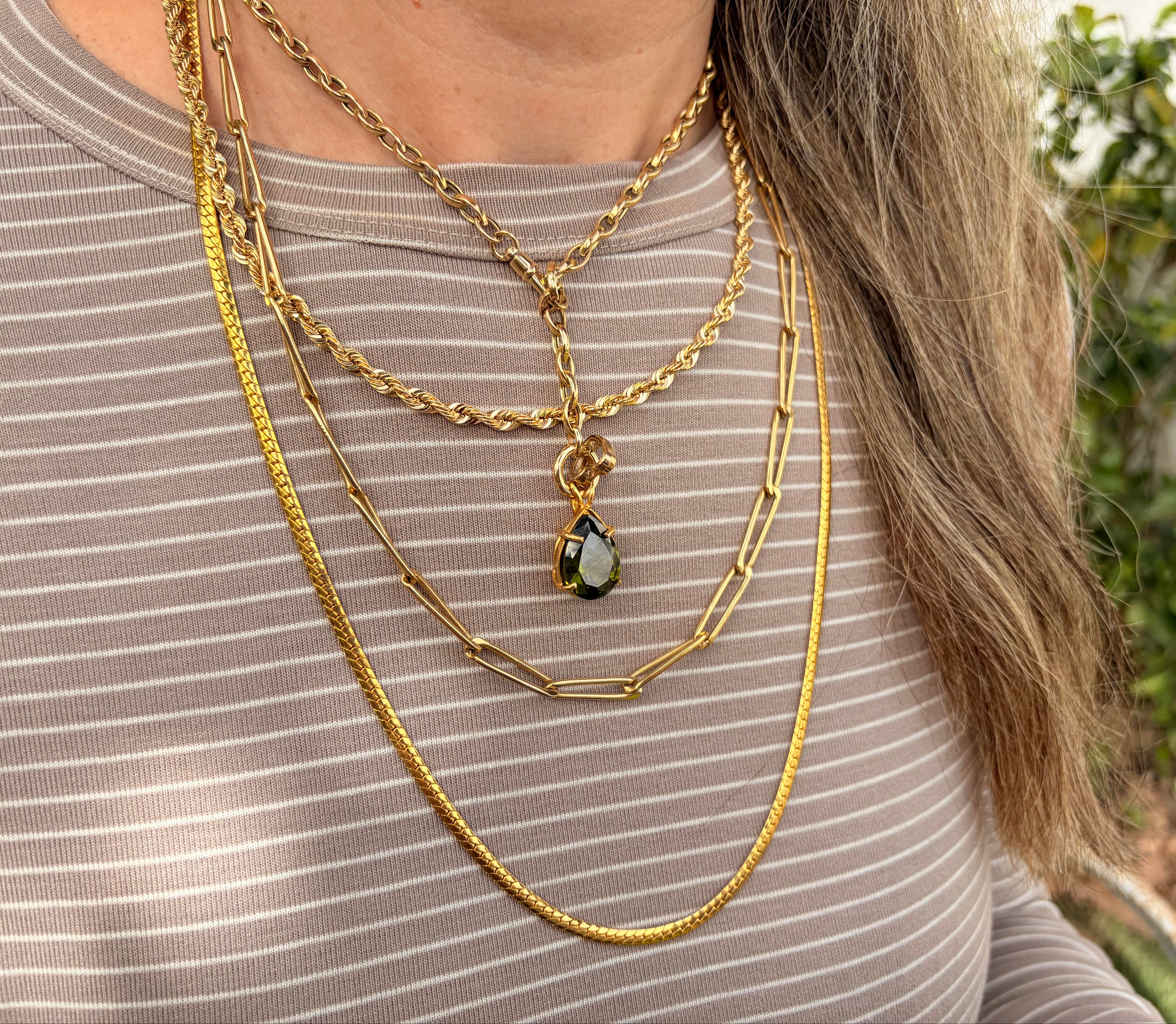 A close-up of a person wearing layered gold necklaces with focus on 14k yellow gold dog clip swivel clasp and round charm holder necklace, including a teardrop pendant with a dark green and blue iridescent stone, over a taupe ribbed top with green foliage in the background.