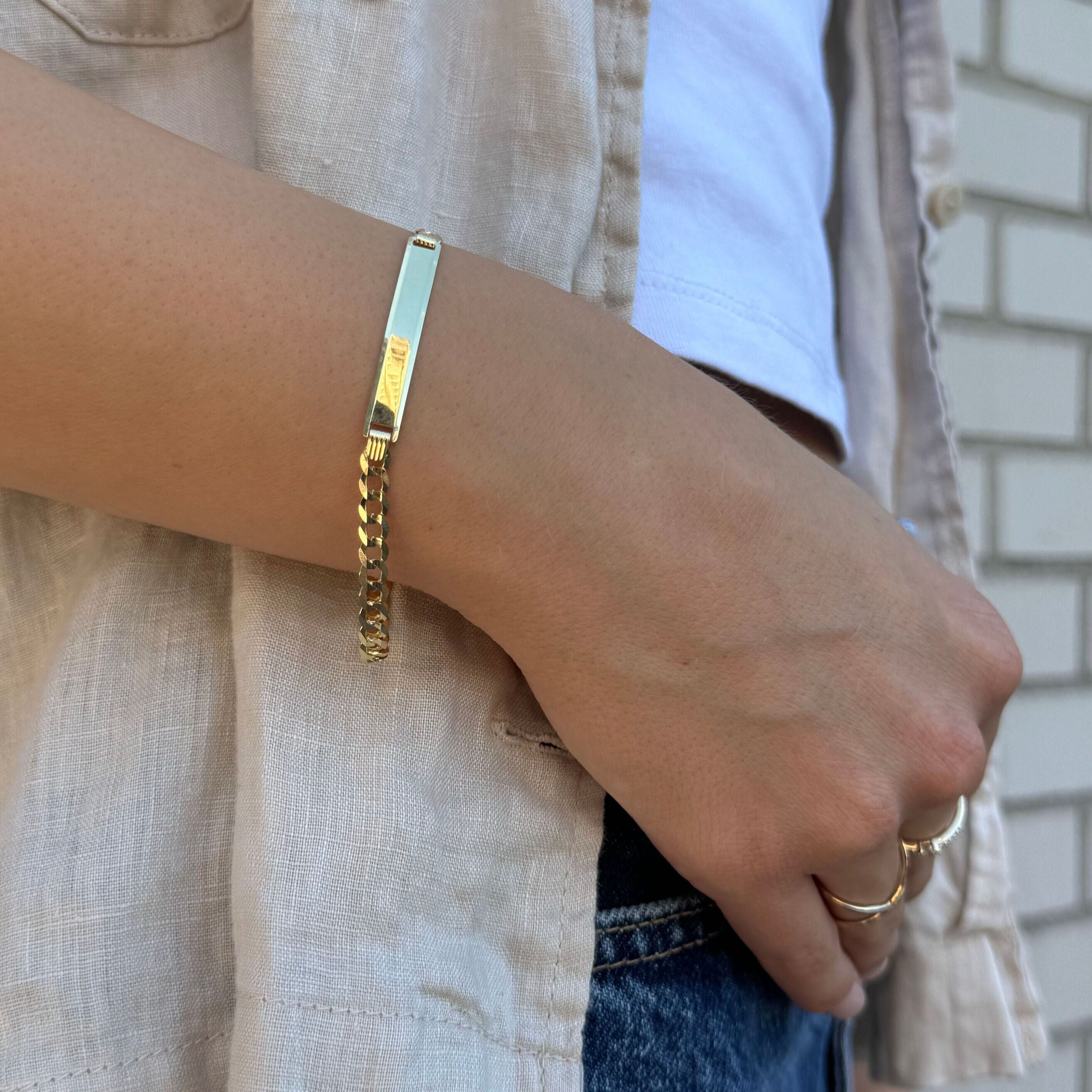 14K solid gold ID bar curb link bracelet styled on the wrist with a cream shirt, white tank, and blue jeans. The polished gold bar adds sleek sophistication and modern versatility, making it ideal for casual outings, everyday wear, or weekend gatherings.